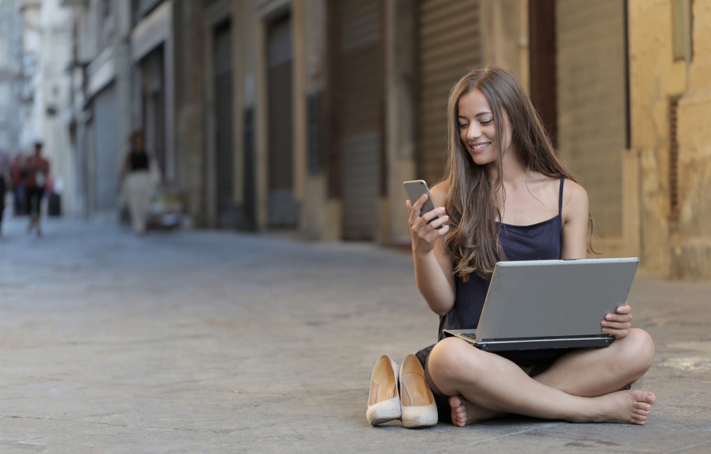 Image of a woman sitting on a floor while using a smartphone and a laptop